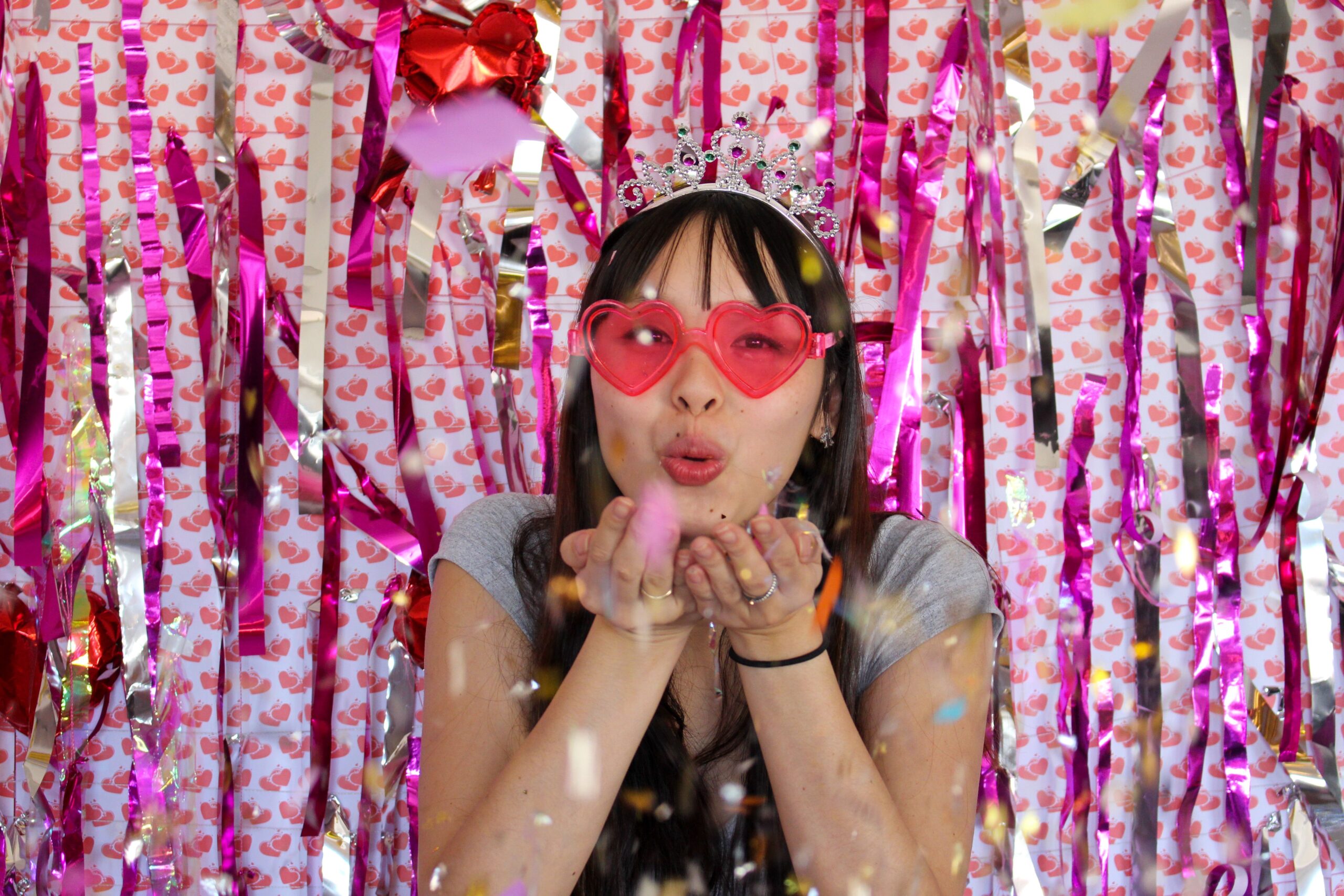 Young woman in heart sunglasses and tiara blowing confetti at a festive party.
